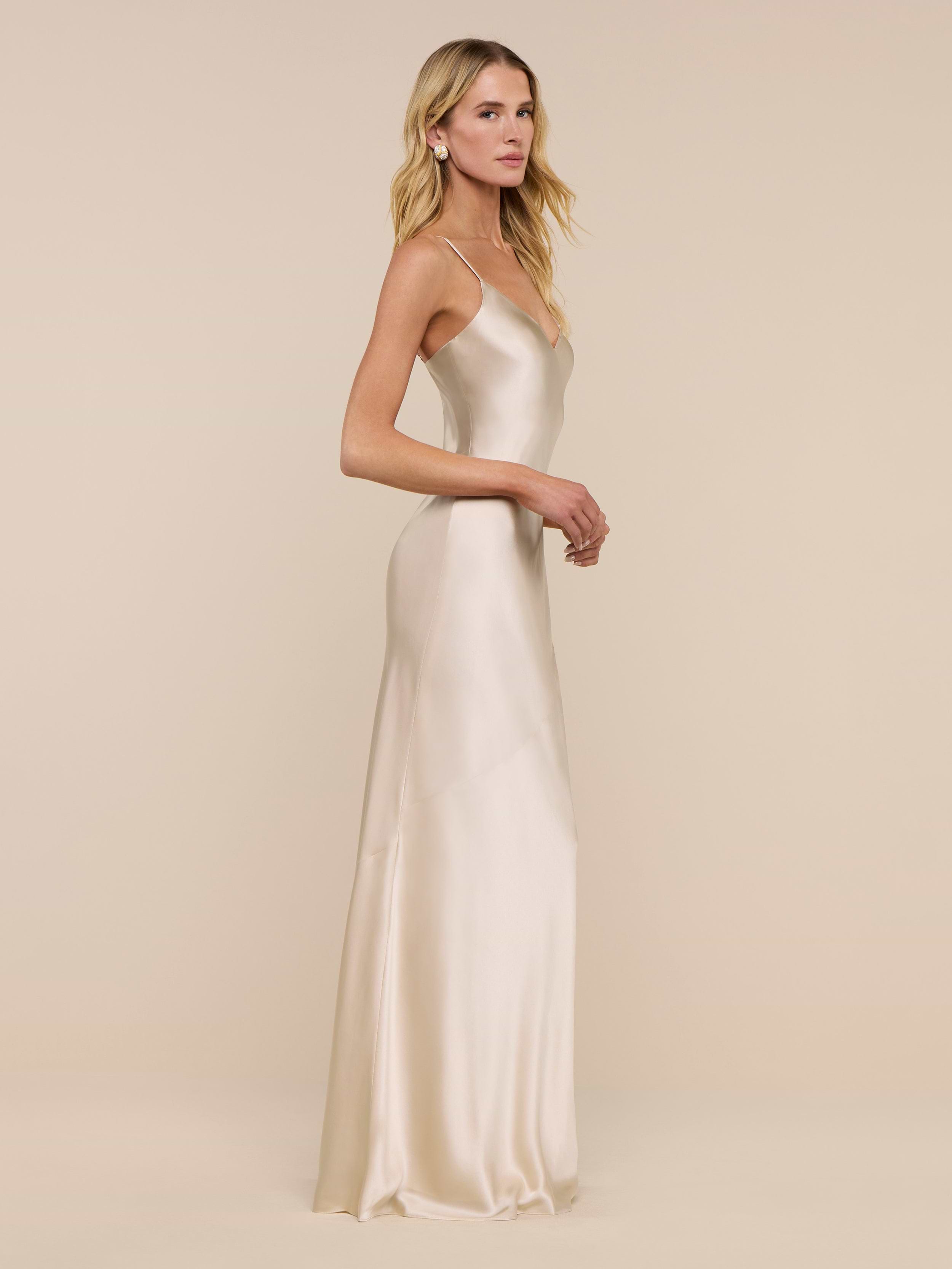 A woman is seen in profile against a plain white background, wearing the Serita Silk Gown—a long, champagne-hued charmeuse dress. Her long blonde hair falls loosely as she looks ahead, arms relaxed at her sides.