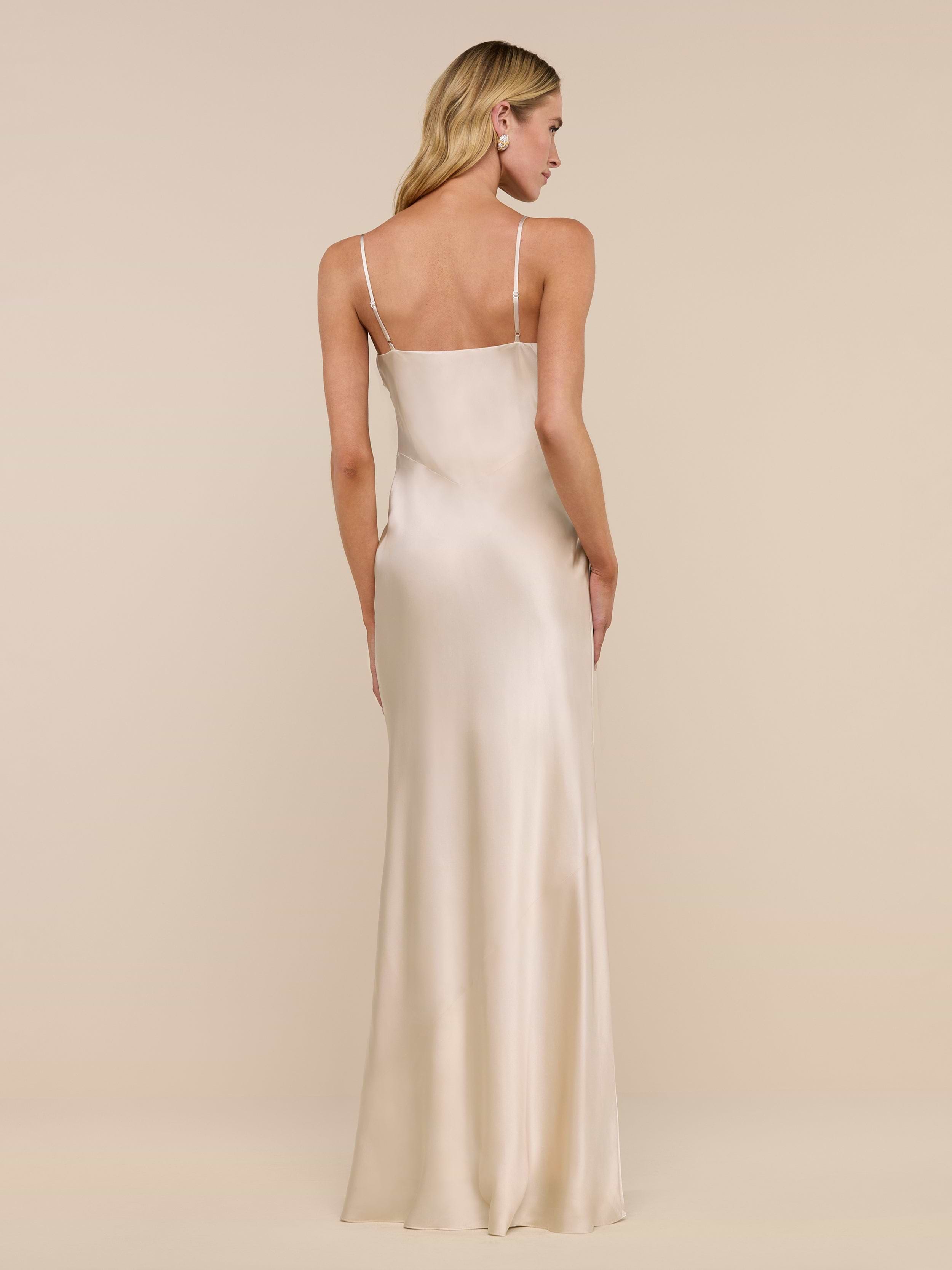 A woman with long blonde hair wears the floor-length Serita Silk Gown in light champagne, featuring thin straps, as she stands facing away against a plain white background.