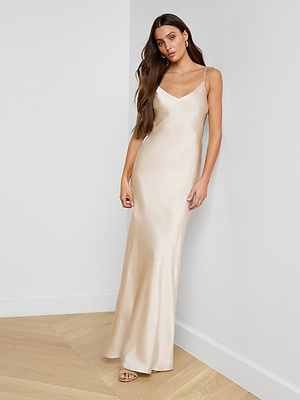 A woman with long brown hair poses against a white wall on a wooden floor, wearing the Serita Silk Gown—a sleeveless champagne slip dress—while looking at the camera.