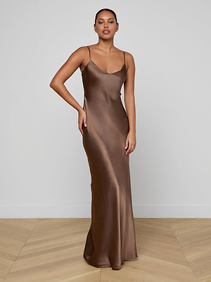 A woman stands on a wooden floor wearing the Serita Silk Gown, a long brown slip dress with thin straps—her hair pulled back as she poses against a plain wall, ideal for an elegant wedding guest look.