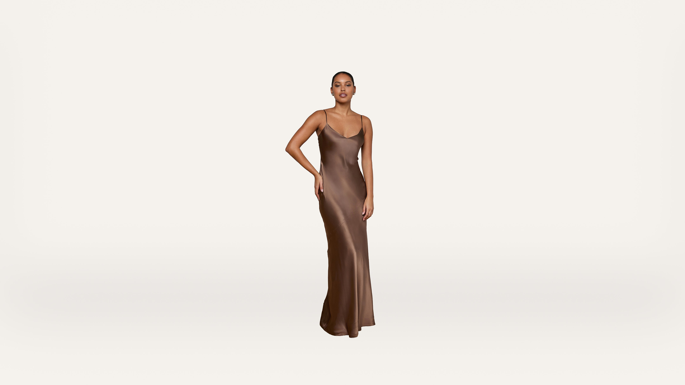 A person stands on a white background wearing a long, sleeveless, bronze satin dress, with one hand resting on their hip and a neutral expression.