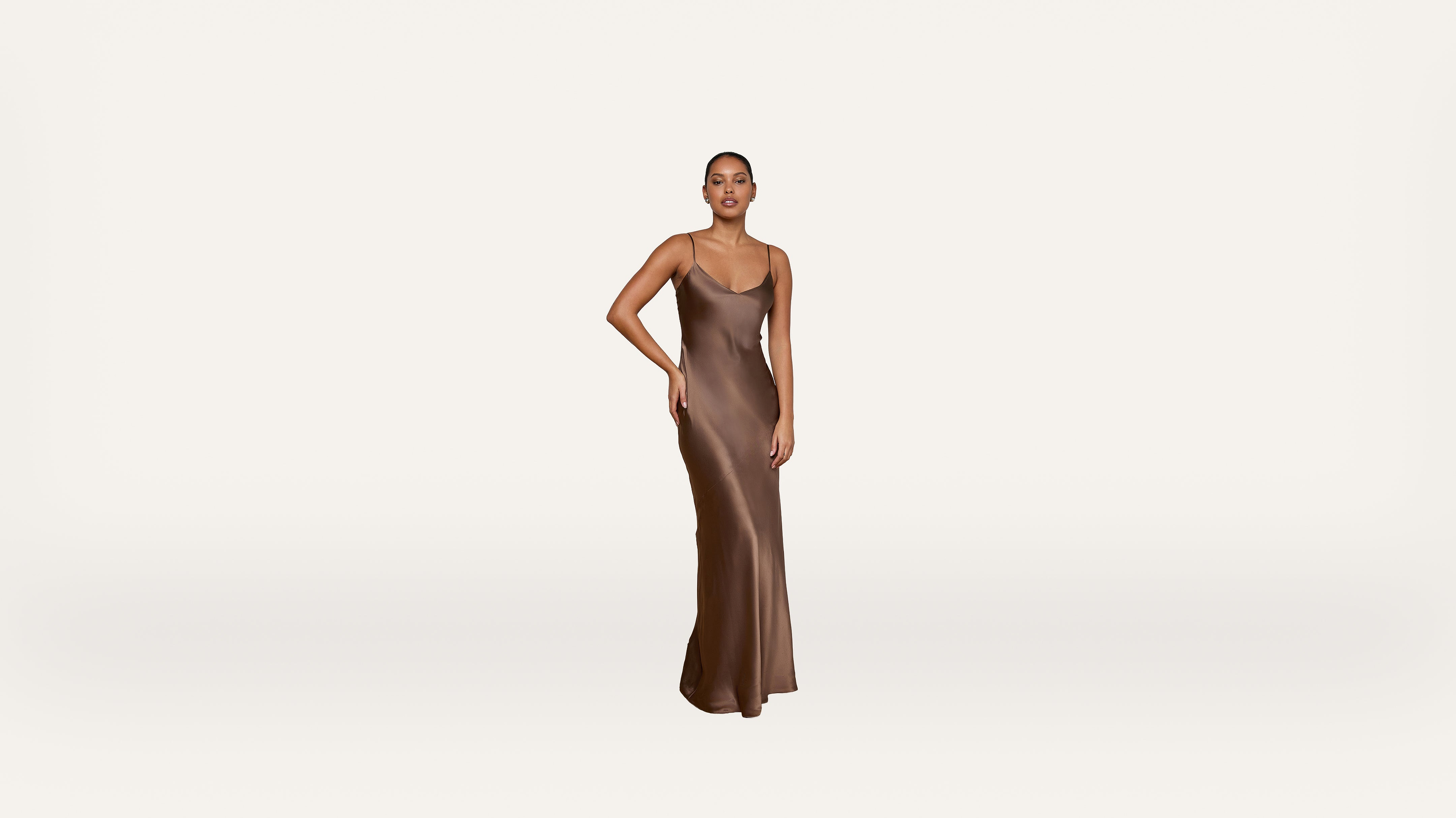 A person stands on a white background wearing a long, sleeveless, bronze satin dress, with one hand resting on their hip and a neutral expression.