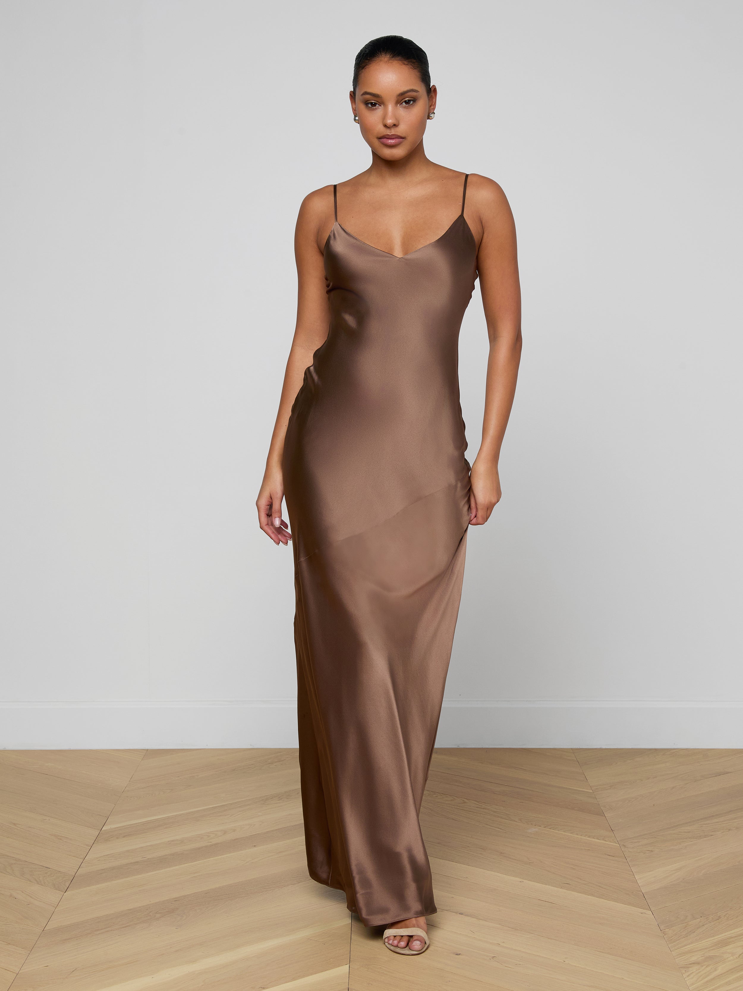 A woman stands indoors on a wooden floor wearing the Serita Silk Gown, a long brown slip dress with thin straps and open-toe heels. The plain, light background highlights this elegant choice for a wedding guest dress.