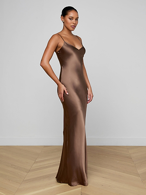 A woman stands in a minimal room wearing the Serita Silk Gown—a long, fitted brown silk dress with thin straps—her hair pulled back and earrings on, posing stylishly on a light wooden floor. Perfect as a chic wedding guest dress.