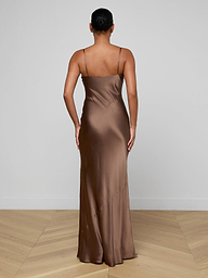 A person stands with their back to the camera, wearing the Serita Silk Gown—a long brown slip dress with thin straps, close fit, and floor-length hem. Their hair is in a neat bun; the setting features light walls and a wooden floor.