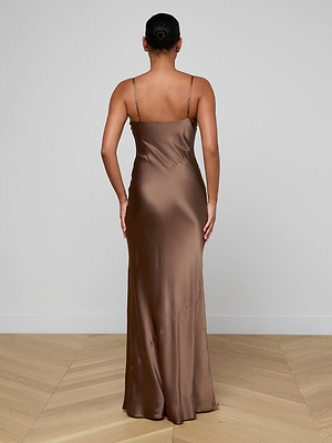 A person stands with their back to the camera, wearing the Serita Silk Gown—a long brown slip dress with thin straps, close fit, and floor-length hem. Their hair is in a neat bun; the setting features light walls and a wooden floor.