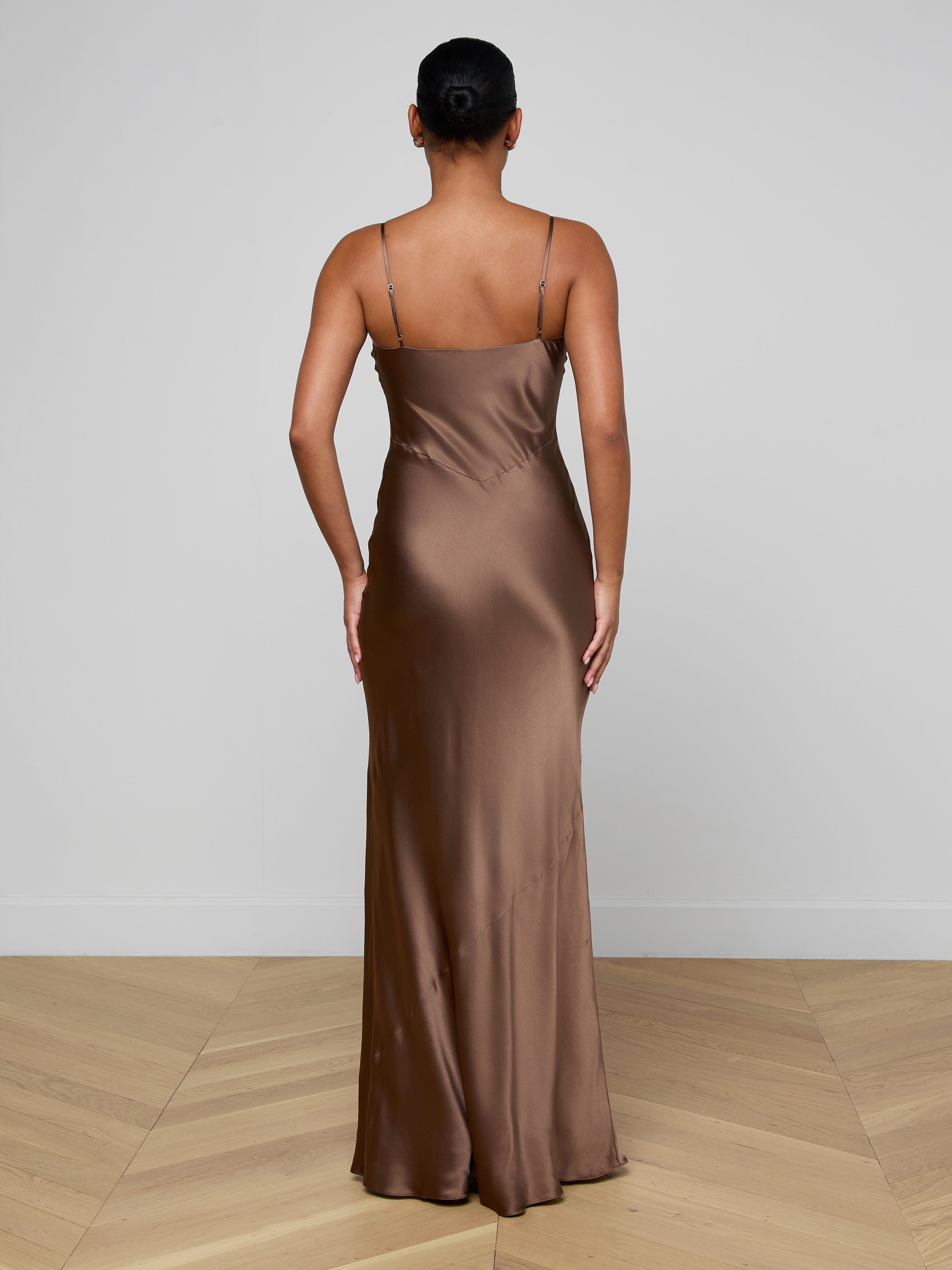 A person stands with their back to the camera, wearing the Serita Silk Gown—a long brown slip dress with thin straps, close fit, and floor-length hem. Their hair is in a neat bun; the setting features light walls and a wooden floor.