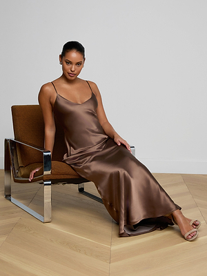 A woman in the Serita Silk Gown, featuring shiny brown silk, sits on a brown chair with metal arms atop a light wood herringbone floor against a white wall. With styled-back hair and sandals, her look is perfect for a wedding guest dress.
