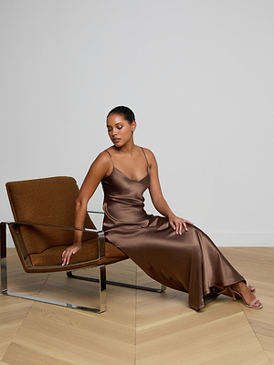 A woman in the Serita Silk Gown sits on a modern chair, one hand on the chair and the other on her knee, against a white wall and wooden floor. This brown slip dress is perfect as a chic wedding guest outfit.