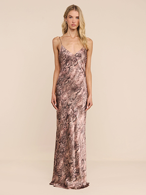 A woman with long blonde hair wears the Serita Silk Gown, a sleeveless, floor-length dress featuring brown and pink marble patterns, thin straps, and a cowl neckline, standing before a plain beige background.