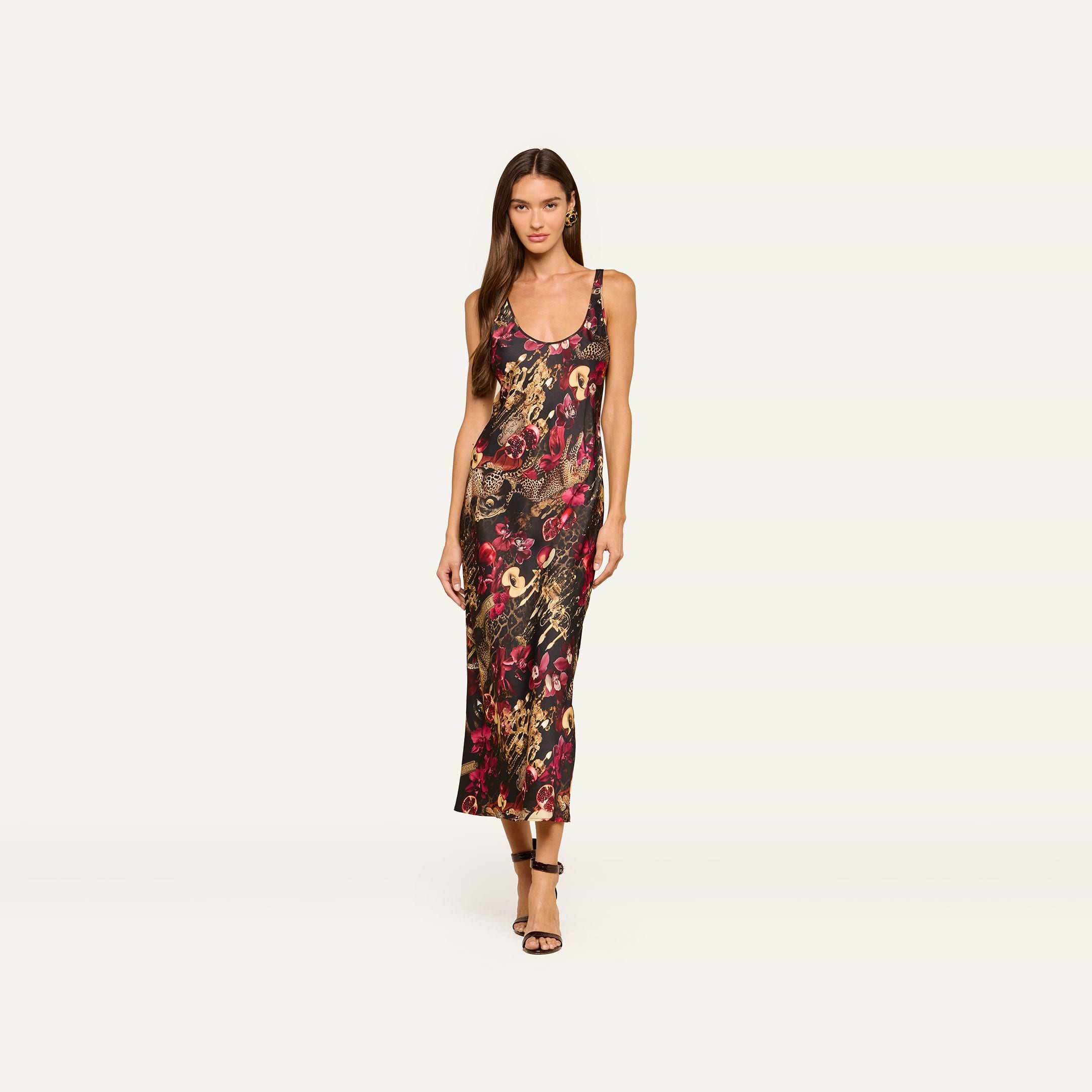 A woman models the Akiya Satin Maxi Dress—a colorful, bias-cut floral print tank dress with thin straps—paired with strappy heels and posed against a plain white background.
