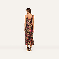 A woman stands facing away, wearing the Akiya Satin Maxi Dress—a sleeveless, ankle-length tank dress with a bold collage print in dark and bright tones. She has long brown hair and is styled with black high-heeled shoes against a plain off-white background.
