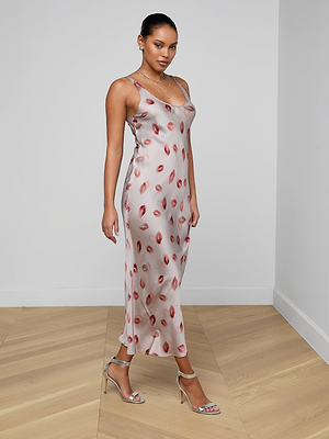 A woman in the Akiya Satin Maxi Dress, featuring a red lips print and bias cut, stands in a minimalist room with light wood floors and white walls. She completes her evening look with silver high-heeled sandals, gazing at the camera.