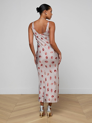 A woman faces away in the Akiya Satin Maxi Dress, a light pink, bias-cut evening gown with red lip prints, paired with silver heels. She poses in a minimal room with a white wall and light wood floor.