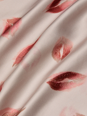 A close-up of the Akiya Satin Maxi Dress shows light pink bias-cut satin scattered with red lipstick kiss marks in various shades and sizes. The fabric is slightly folded, perfect for an evening look, with soft shadows enhancing its elegance.