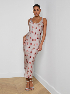 A woman stands against a white wall wearing the Akiya Satin Maxi Dress, a sleeveless ankle-length bias-cut satin dress with a pink base and red lip print. She has her hair tied back and pairs it with silver heels and layered necklaces.