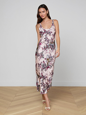 A woman with long brown hair wears the Akiya Satin Maxi Dress, a sleeveless, ankle-length evening piece featuring a pink base with vibrant floral and butterfly prints. She stands on a wooden floor against a plain light gray wall, facing the camera.