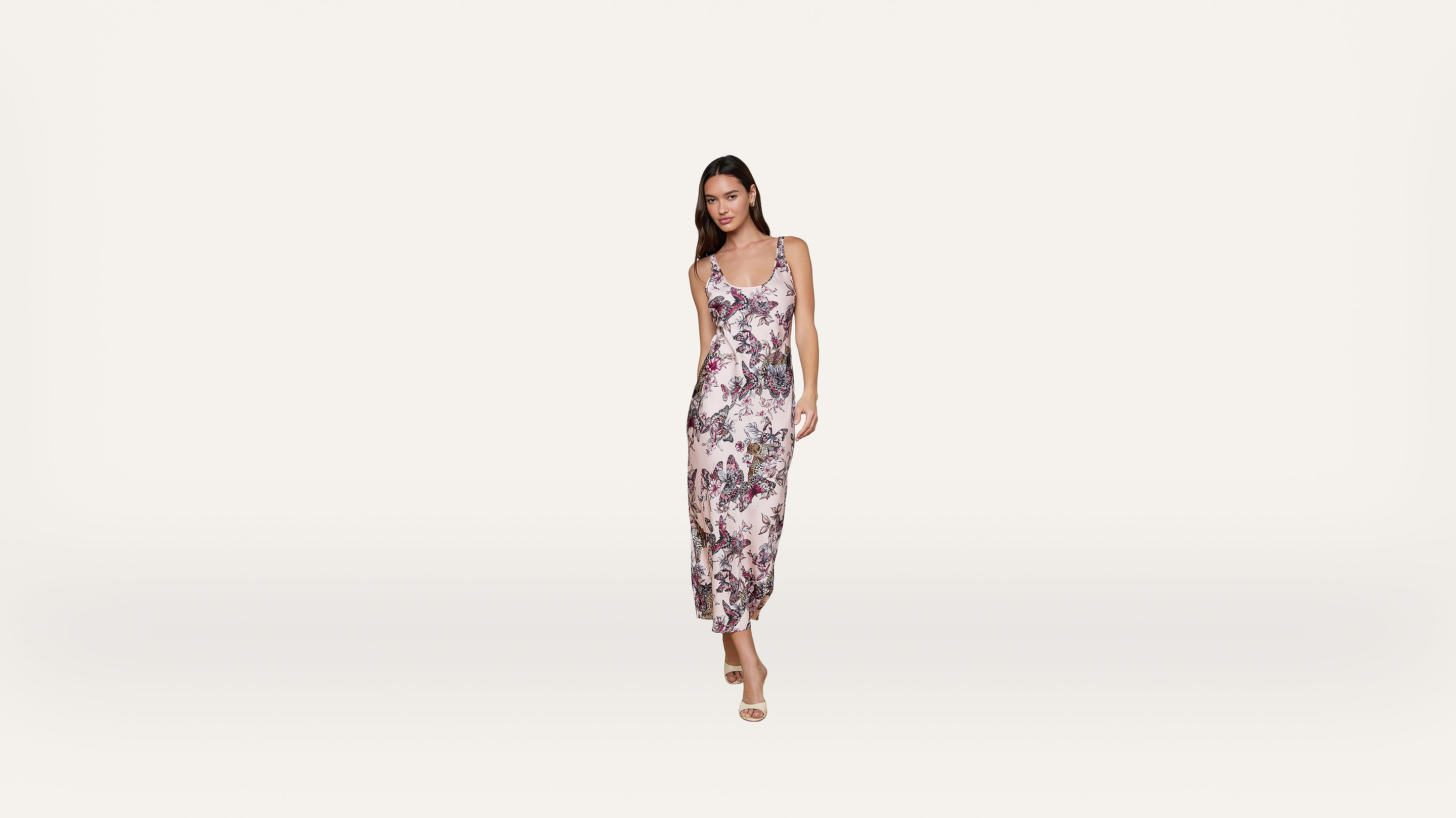 Woman wearing a sleeveless, floral-patterned midi dress and sandals stands against a plain white background.