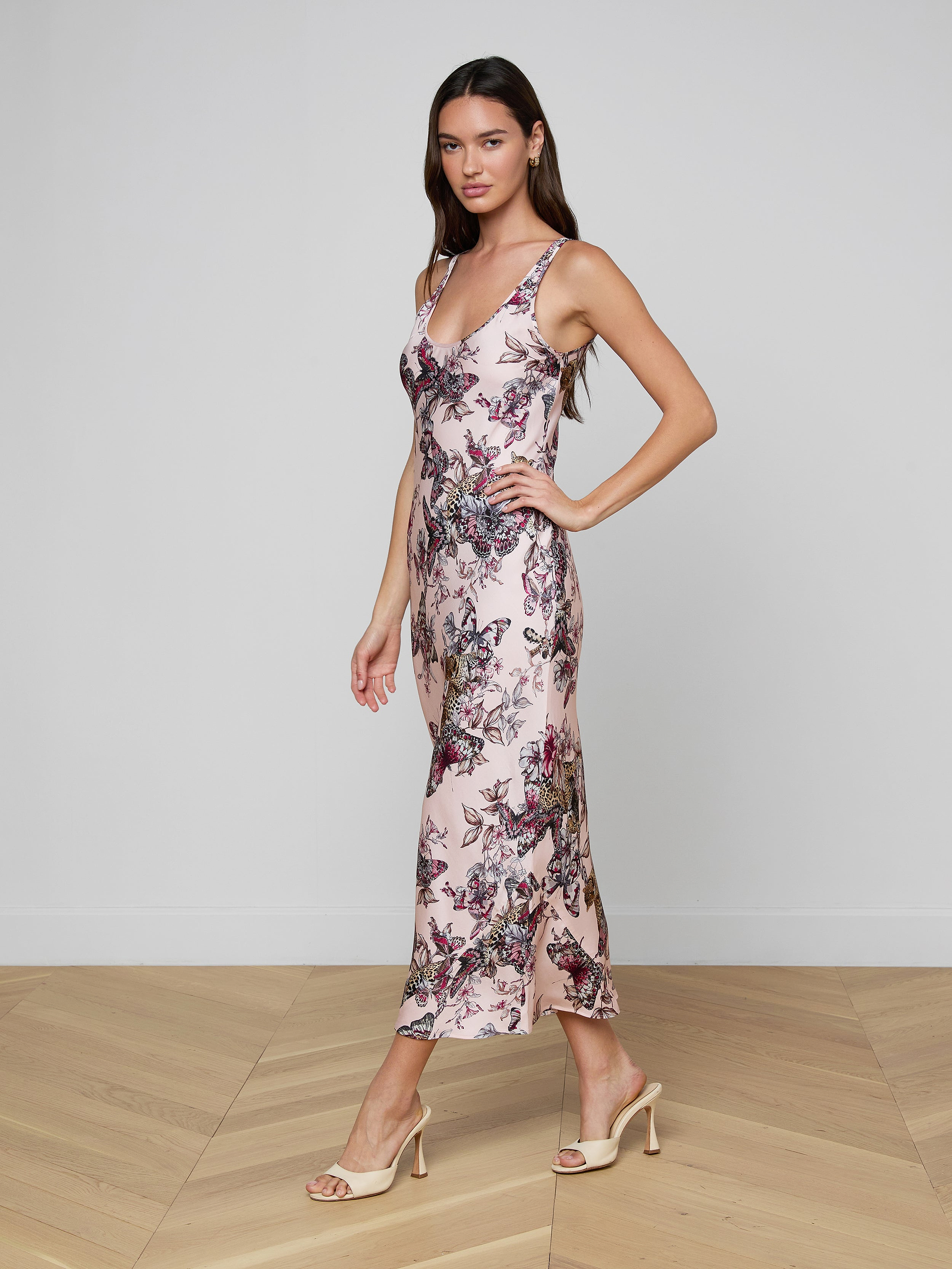 A woman stands in profile against a plain off-white background, wearing the Akiya Satin Maxi Dress—an ankle-length, sleeveless bias-cut dress featuring a butterfly jungle print—paired with beige heels.