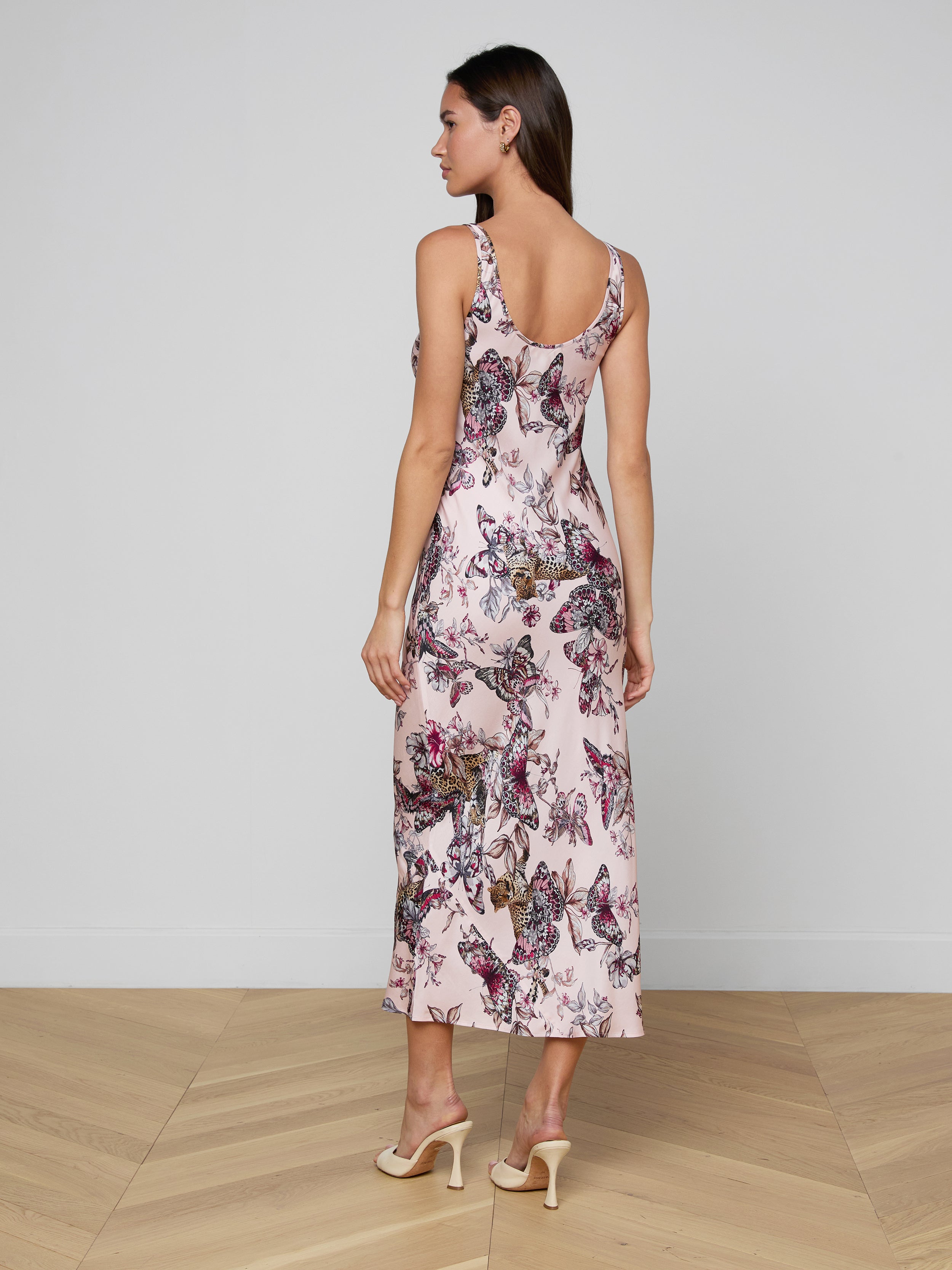 A woman stands facing away against a plain, light background, wearing the Akiya Satin Maxi Dress—a bias-cut satin gown featuring a butterfly jungle print—paired with light-colored high heels.