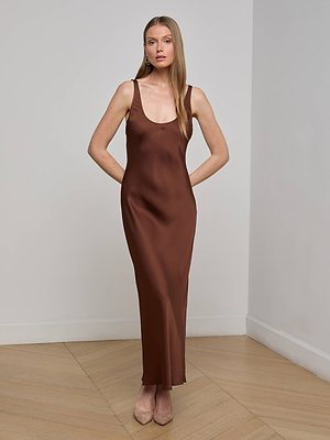 A woman stands indoors in the Akiya Satin Maxi Dress, a sleeveless, brown, ankle-length bias-cut slip. She pairs it with beige heels, her long hair straight and arms relaxed at her sides against a plain wall.