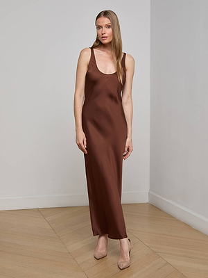 A woman stands on a wooden floor indoors, wearing the Akiya Satin Maxi Dress in brown with beige heels. She has long straight hair, a neutral expression, and one hand by her side.