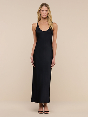 A woman with long blonde hair stands facing forward in the Akiya Satin Maxi Dress, a long sleeveless black dress with a scoop neckline, styled with black heels against a beige background. She poses simply and relaxed.