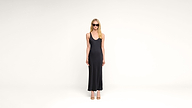 A woman with long blonde hair stands against a plain white background, wearing a black sleeveless dress, sunglasses, and open-toed heels.