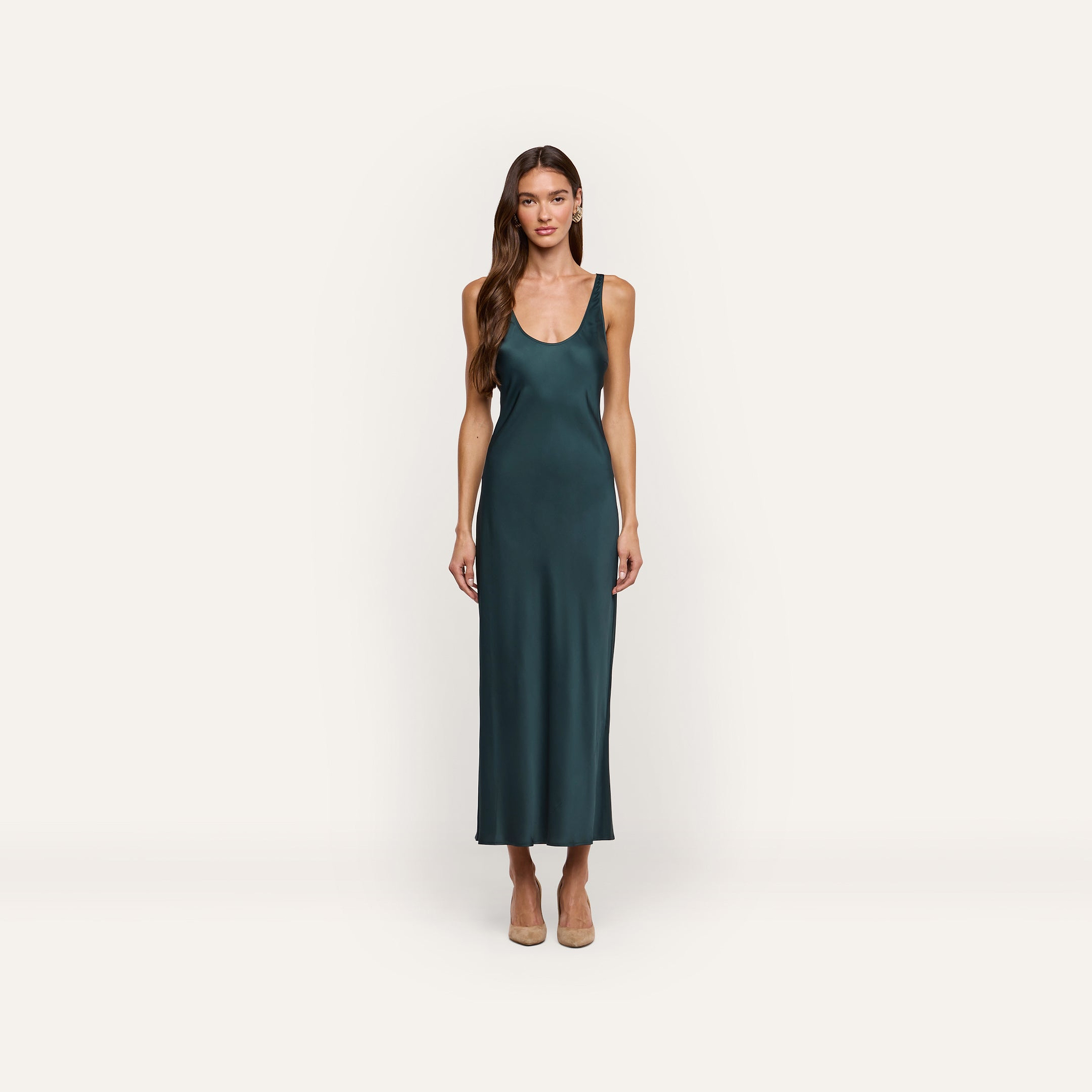A woman wearing an Akiya Satin Maxi Dress in forest green, featuring a bias-cut, ankle-length scoop-neck style paired with tan pumps.