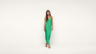 A woman stands against a plain white background wearing a sleeveless, bright green, floor-length dress and beige open-toe heels.