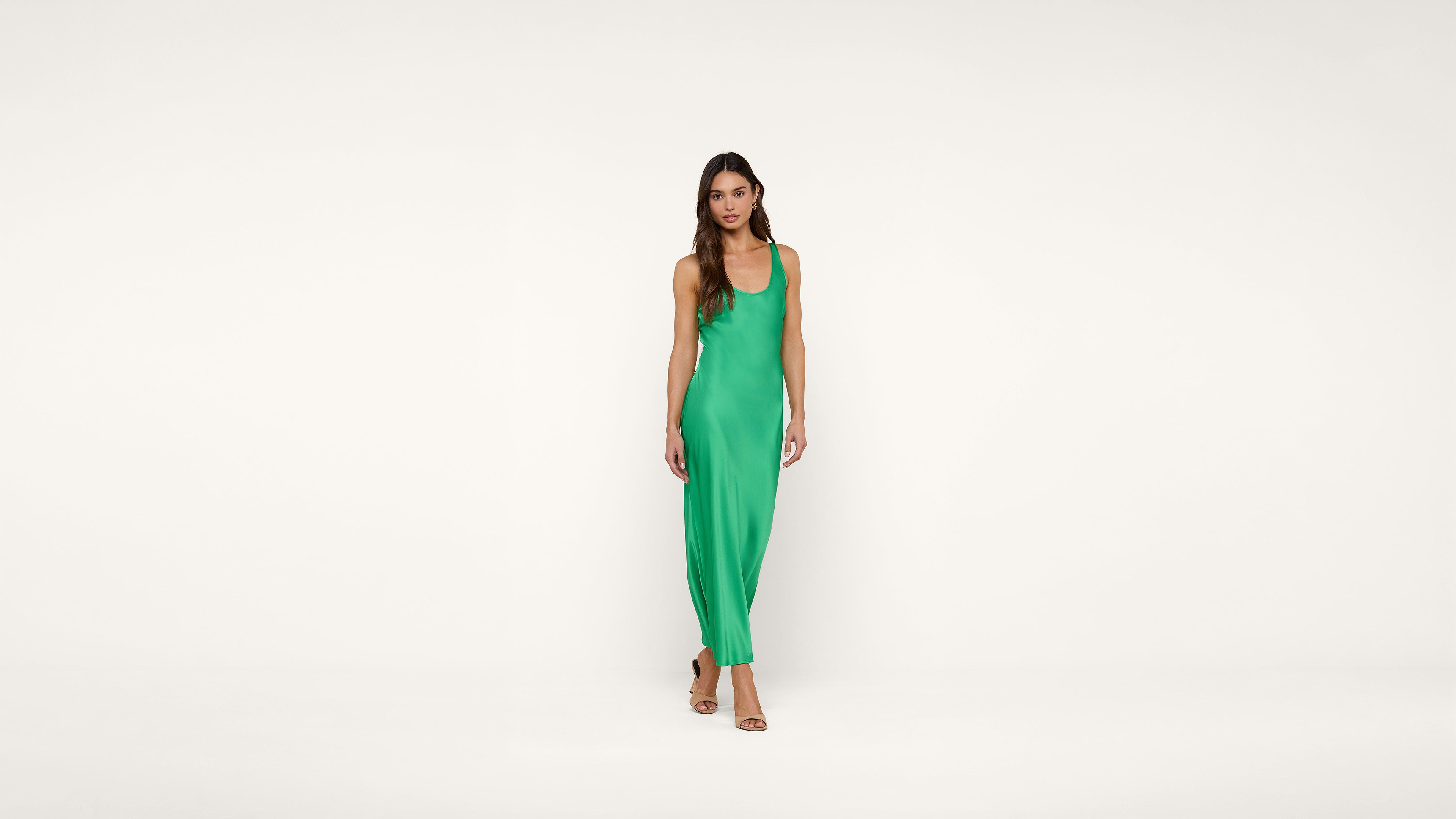 A woman stands against a plain white background wearing a sleeveless, bright green, floor-length dress and beige open-toe heels.