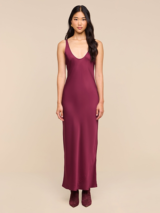 A woman wearing the Akiya Satin Maxi Dress—a sleeveless, burgundy satin dress with a scoop neckline—stands facing forward against a beige background.