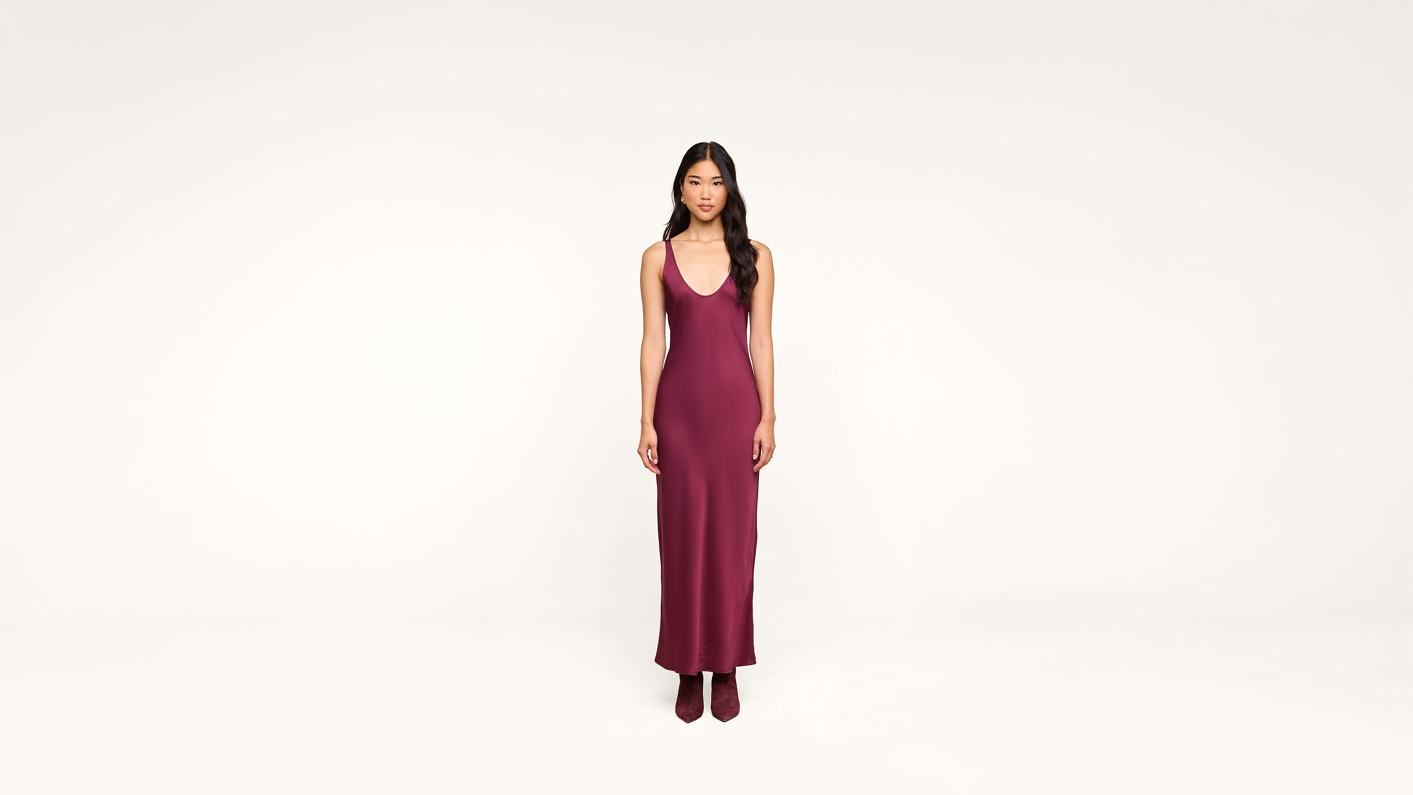 A woman with long dark hair stands facing forward, wearing a sleeveless, deep burgundy maxi dress and matching boots against a plain white background.