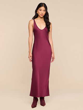 A woman stands against a beige background wearing the Akiya Satin Maxi Dress in deep burgundy, styled as a flowing sleeveless tank dress and paired with matching ankle boots. Her loose, wavy hair completes the elegant look.