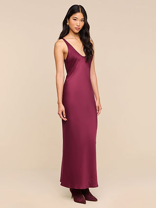A woman stands against a plain beige background, looking at the camera with a relaxed expression, wearing the Akiya Satin Maxi Dress—an elegant sleeveless tank dress in deep red satin—and matching boots.
