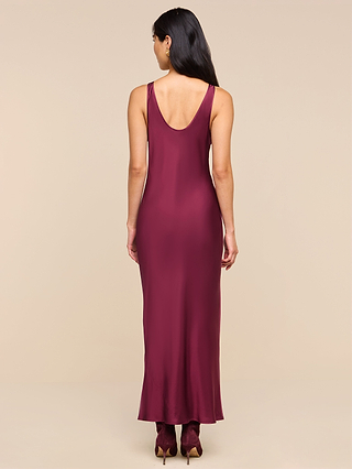 A woman with long dark hair stands facing away against a light beige background, wearing the Akiya Satin Maxi Dress—a sleeveless, deep burgundy gown in satin with a scoop back. She completes her look with matching heels.
