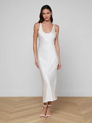 A woman stands on a wooden floor against a white wall, wearing the Akiya Satin Maxi Dress—a long, sleeveless tank dress with a scoopneck—and white strappy heels. She faces the camera with a neutral expression, arms relaxed at her sides.