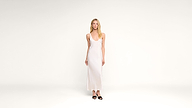 A woman wearing a long, sleeveless white dress and black sandals stands against a plain white background, looking forward with a neutral expression.