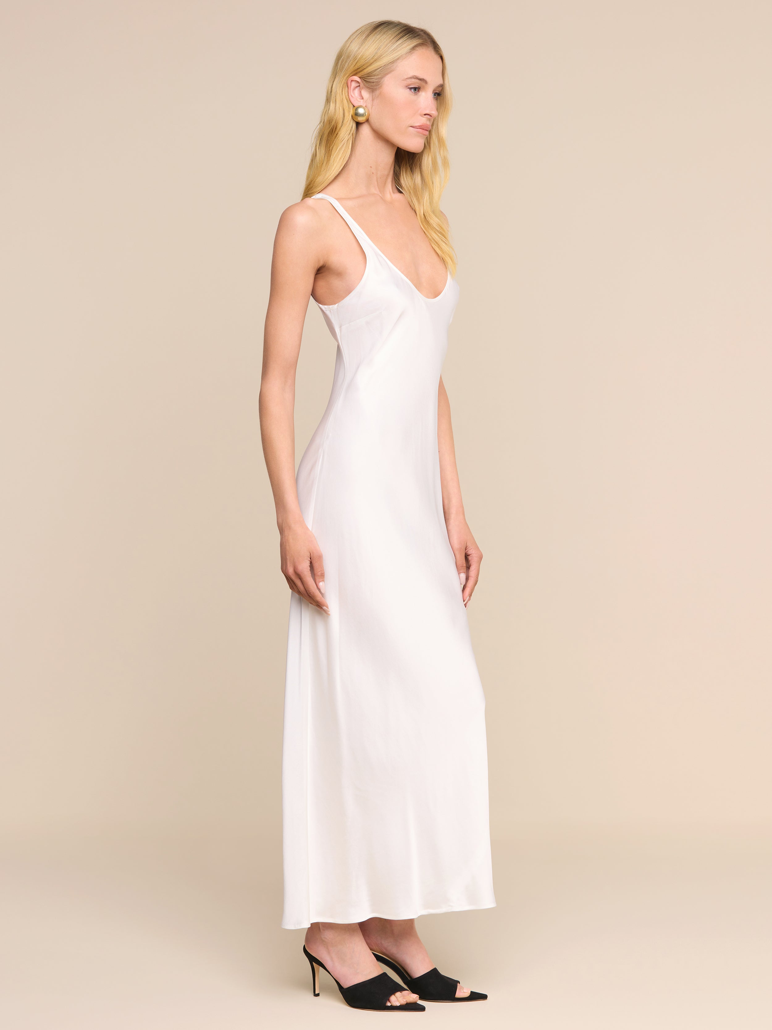 A woman wearing the Akiya Satin Maxi Dress—a long, light-colored bias-cut tank dress with a scoop neck—paired with black high-heeled shoes stands sideways against a plain white background.