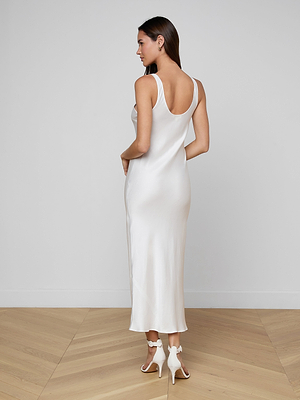 A woman stands in a minimal room on wooden flooring, facing away from the camera, wearing the Akiya Satin Maxi Dress and white high-heeled sandals.
