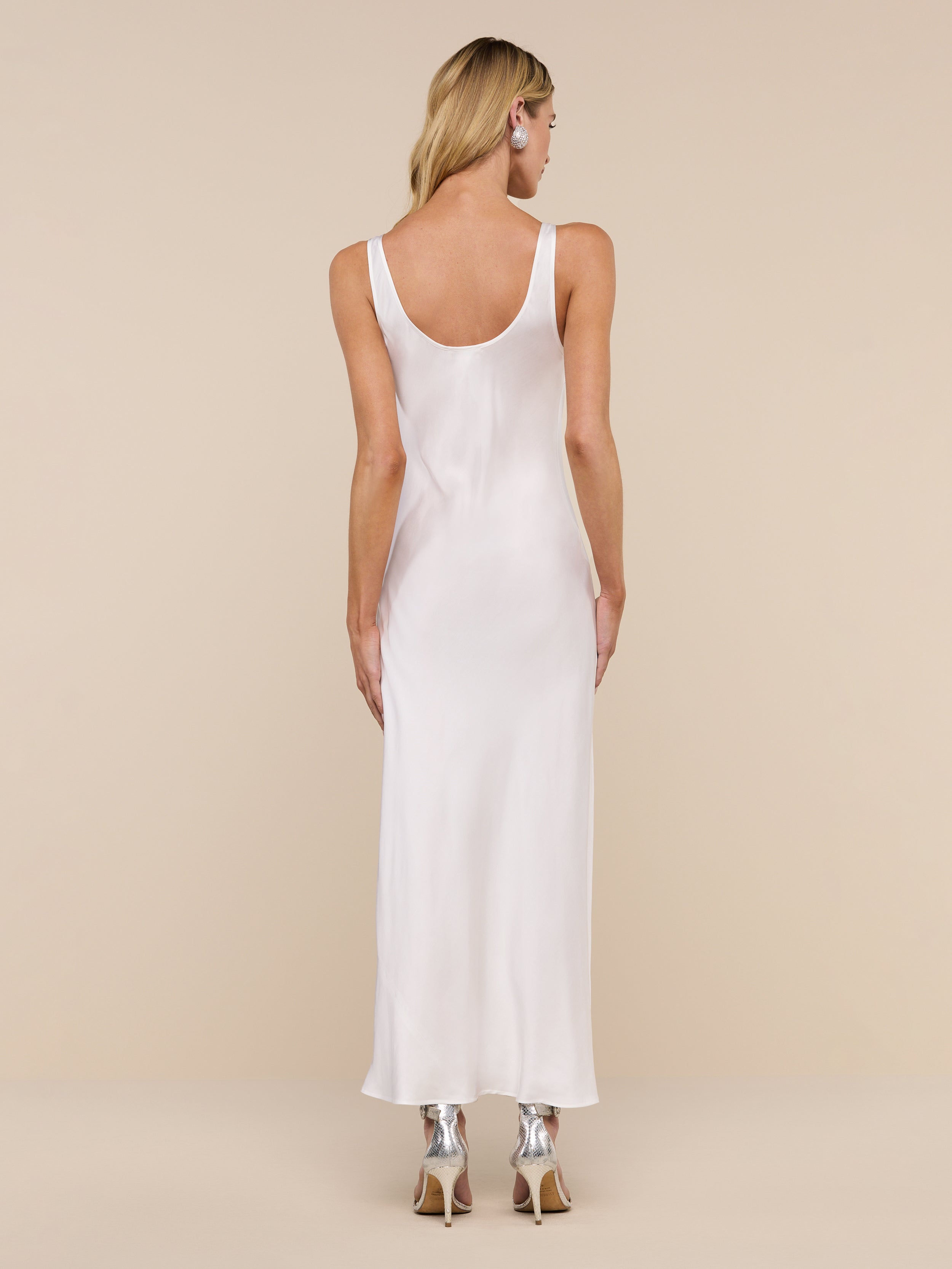 A woman with long blonde hair, wearing the Akiya Satin Maxi Dress and black heels, stands facing away from the camera against a plain white background.