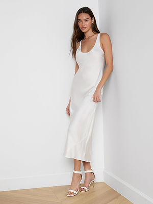 A woman with long brown hair stands in a corner against a white wall and wooden floor, wearing the Akiya Satin Maxi Dress—a sleeveless white satin gown styled elegantly with white high-heeled sandals.