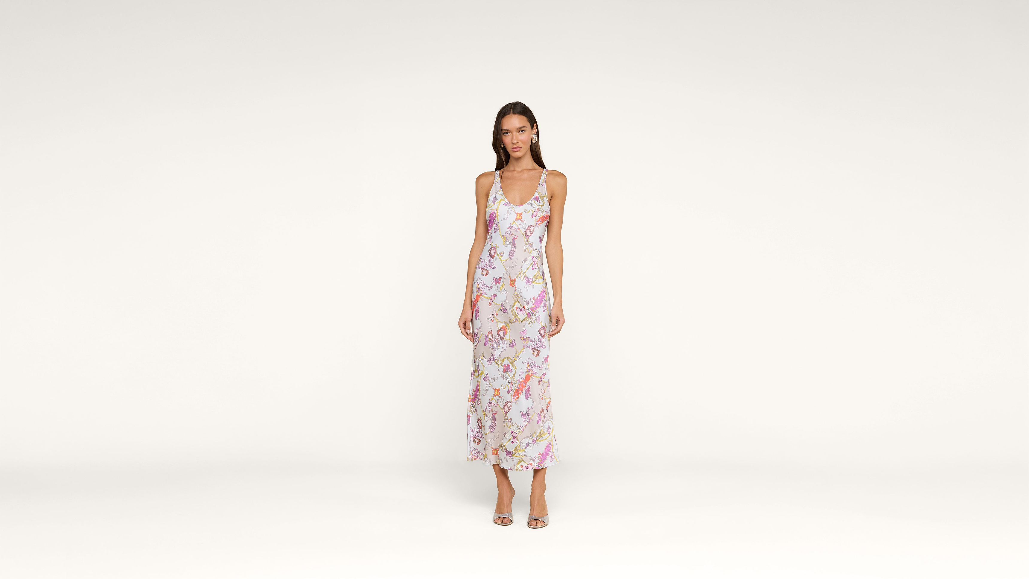 Woman standing against a plain white background, wearing a long, sleeveless, floral dress with open-toe heels. She is facing forward with her arms relaxed at her sides.