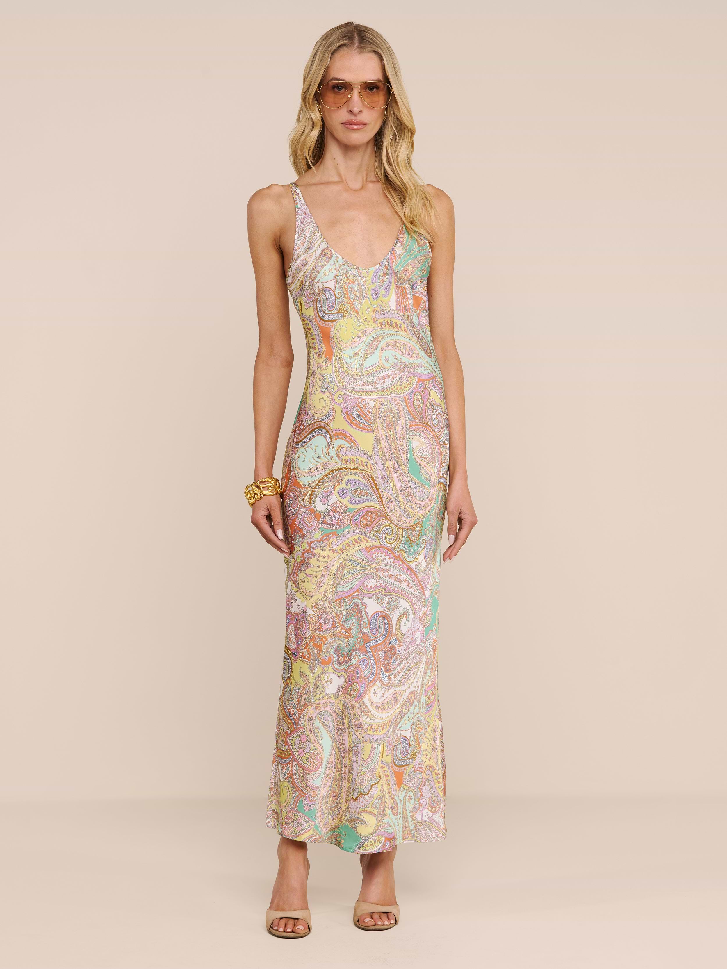 Une femme portant des lunettes de soleil et une longue robe longue sans manches « Akiya Satin Maxi Dress » se tient debout devant un fond beige. Ses cheveux blonds ondulés, son bracelet imposant et ses escarpins à bout ouvert composent une tenue chic, idéale aussi bien pour la journée que pour la soirée.
