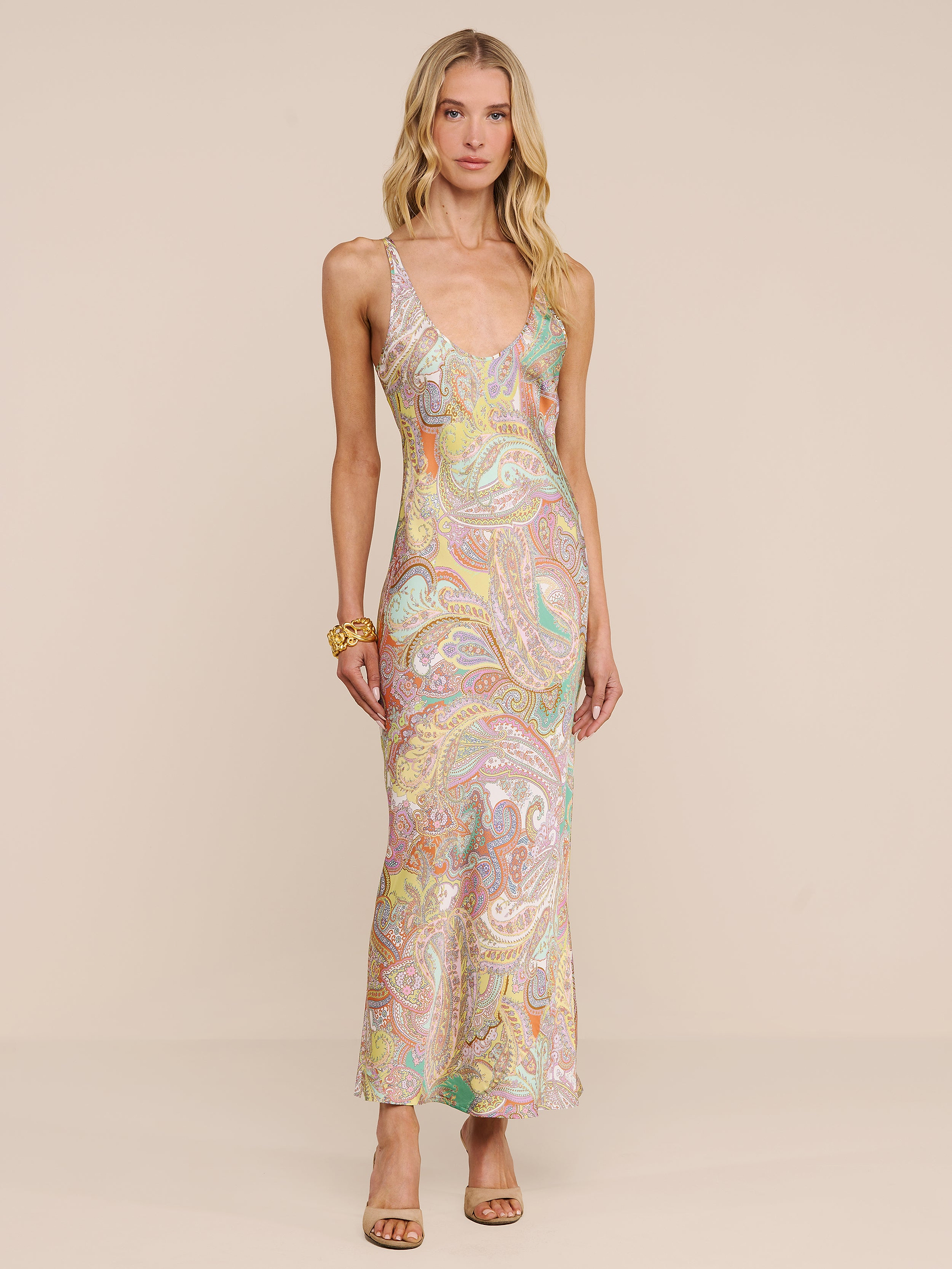 Una donna indossa labito lungo in raso Akiya, un modello senza maniche che arriva fino a terra con una stampa paisley dai toni pastello, in piedi su uno sfondo beige. Completa il suo look con scarpe con tacco aperte in punta e un braccialetto dorato.