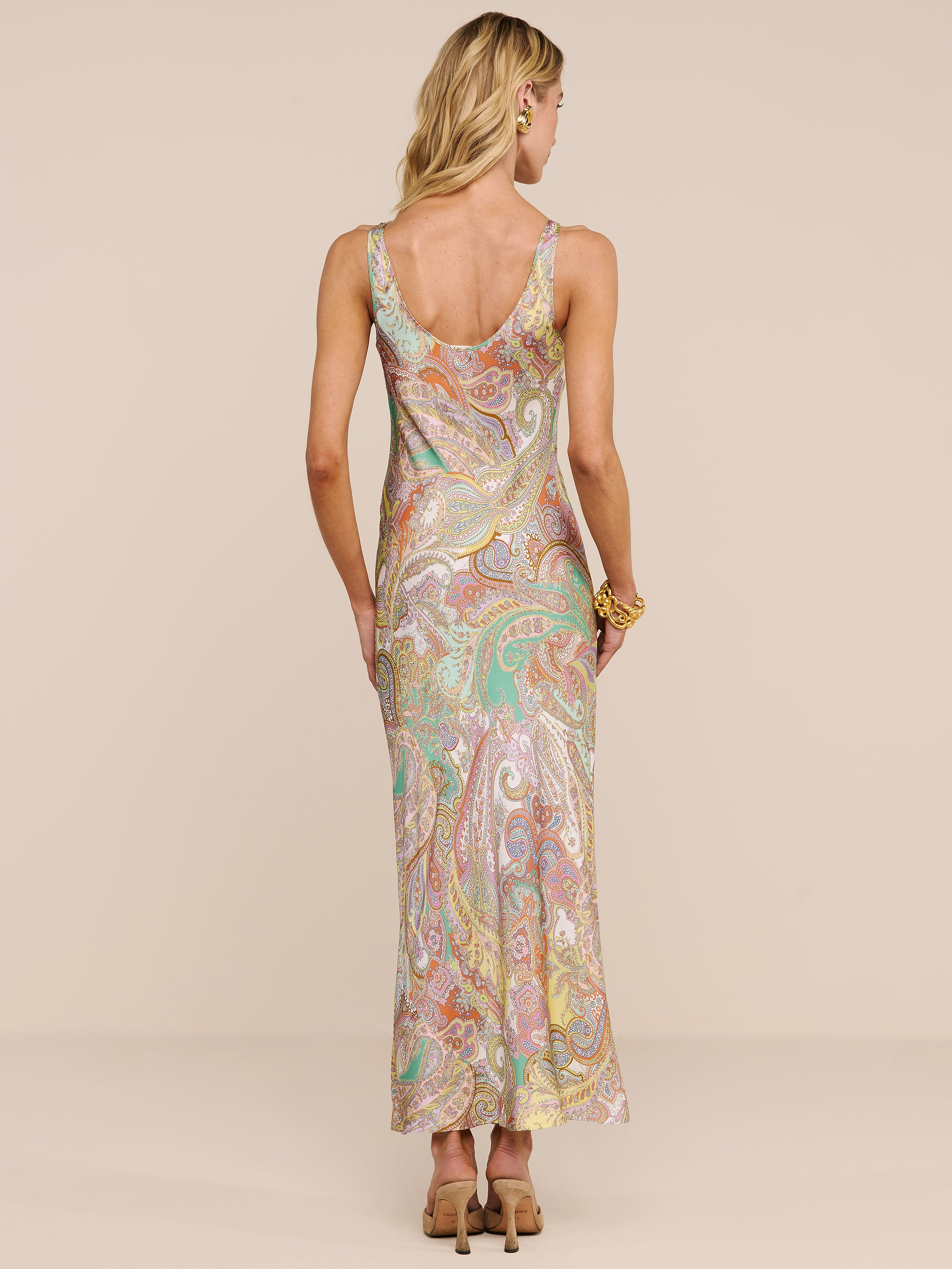 Une femme aux cheveux blonds ondulés, une fleur derrière loreille, est de dos, vêtue de la robe longue en satin « Akiya », un modèle sans manches orné dun imprimé paisley aux teintes pastel. Elle porte des talons et un bracelet, sur un fond beige uni.