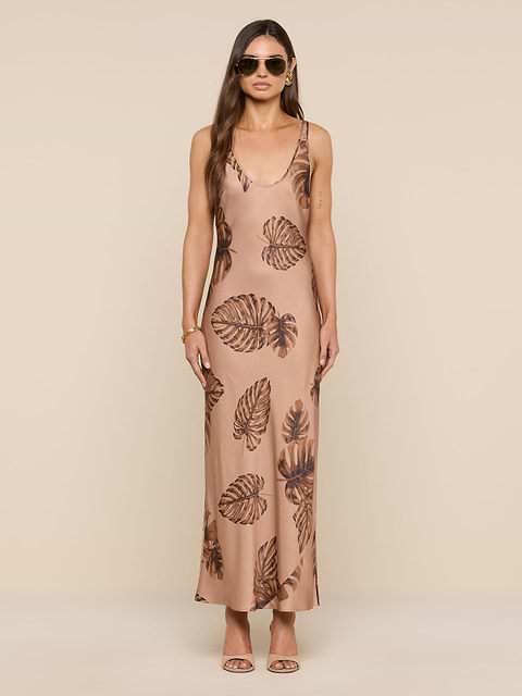 A woman wearing sunglasses and a long, sleeveless tan Akiya Satin Maxi Dress with a brown palm leaf print stands facing forward against a plain white background.