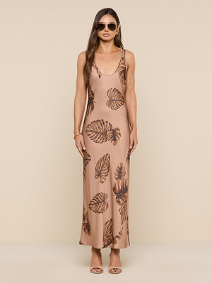 A woman in the Akiya Satin Maxi Dress, featuring a sleeveless, bias-cut design with a large brown palm leaf print, stands against a plain beige background. She accessorizes with sunglasses and open-toed heels, her long hair loose.