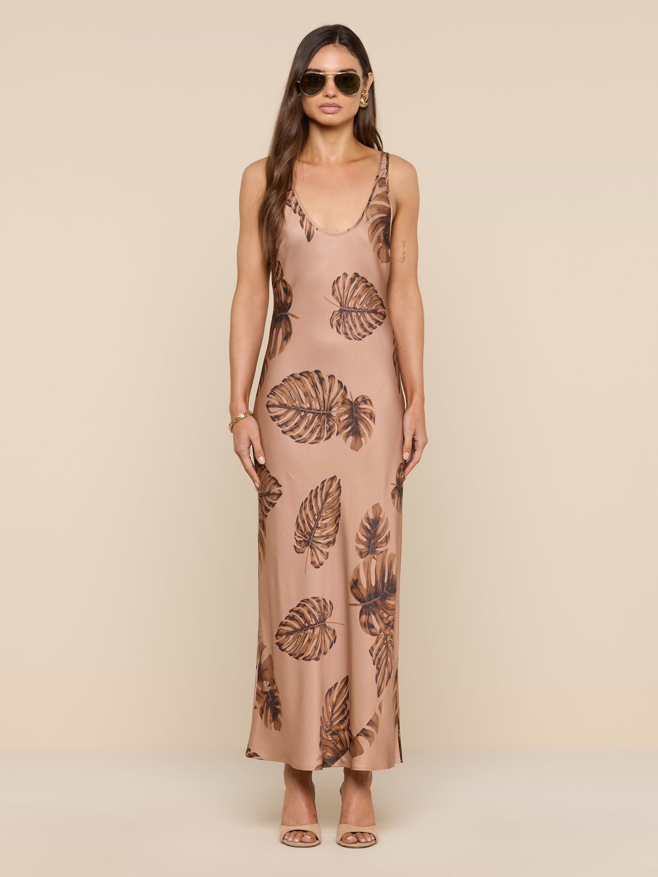 A woman wearing sunglasses and a long, sleeveless tan Akiya Satin Maxi Dress with a brown palm leaf print stands facing forward against a plain white background.