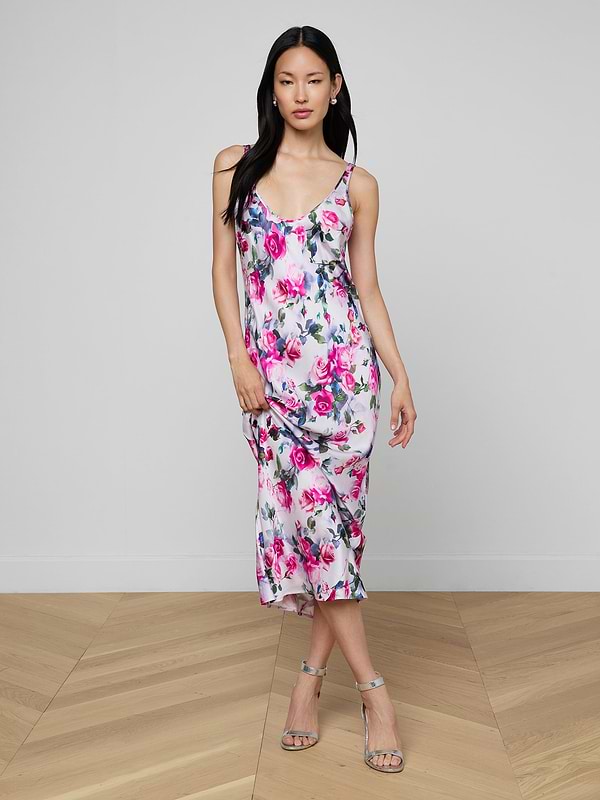 A woman with long dark hair poses gracefully indoors on a wooden floor, wearing the Akiya Satin Maxi Dress in a white and pink rose print with silver strappy heels, one hand holding the hem of her bias-cut satin dress.